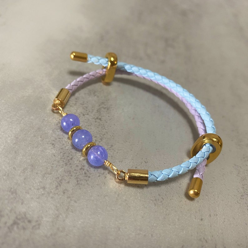 May include: A braided leather bracelet with a light blue and purple colour gradient. The bracelet has three purple beads and gold accents.