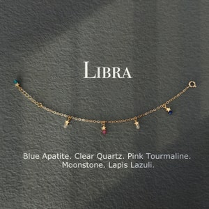 May include: Gold chain bracelet with a star charm and different colored gemstones, including blue apatite, clear quartz, pink tourmaline, moonstone, and lapis lazuli. The bracelet is labeled "LIBRA".