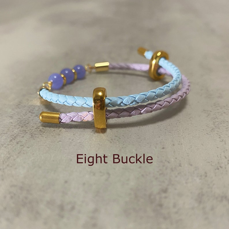 May include: A light blue and lavender braided leather bracelet with a gold buckle and lavender beads. The bracelet is called "Eight Buckle".