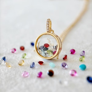 Può includere: Una collana con un medaglione rotondo e trasparente color oro riempito di pietre preziose colorate. Il ciondolo è impreziosito da piccole pietre trasparenti e pende da una delicata catena d'oro. Varie pietre preziose sciolte di diversi colori sono sparse.
