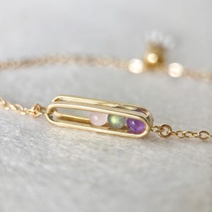 Könnte beinhalten: Ein filigranes goldenes Armband mit einem rechteckigen Anhänger, der drei kleine Edelsteine in Rosa, Grün und Lila enthält. Das Armband hat eine feine Kette und ein schlichtes, elegantes Design.