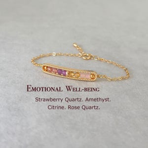 May include: Gold bracelet with a bar charm featuring small pink, purple, and yellow gemstones. The text "EMOTIONAL WELL-BEING" is below the bracelet, followed by "Strawberry Quartz. Amethyst. Citrine. Rose Quartz."