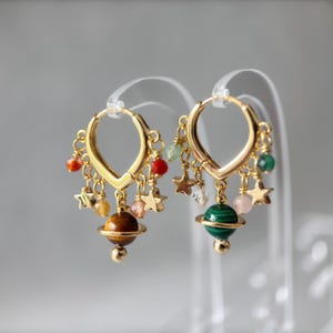 Puede incluir: Pendientes de aro dorados con abalorios celestiales. Cada pendiente presenta un aro dorado con abalorios de estrella colgantes, cuentas de colores y una cuenta en forma de planeta. Una cuenta de planeta es marrón, la otra verde.