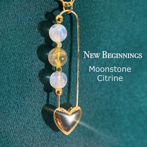 May include: Gold-colored necklace with a heart pendant and three gemstone beads. The beads are a mix of moonstone and citrine. The necklace has the words "New Beginnings" and "Moonstone Citrine" in white text.