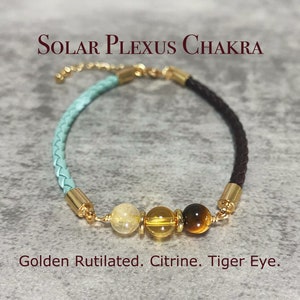 May include: A light blue and brown leather bracelet with gold accents and three gemstone beads: golden rutilated, citrine, and tiger eye. The bracelet is labelled "Solar Plexus Chakra".
