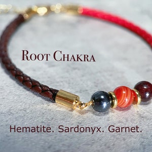 May include: A braided leather bracelet with gold accents and a red cord. The bracelet features beads of hematite, sardonyx, and garnet. Text on the image reads "Root Chakra" and "Hematite. Sardonyx. Garnet."