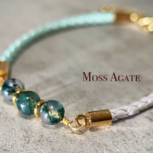 May include: A braided bracelet with moss agate beads and gold-coloured details. The bracelet has a white and light green braided leather band. The text "Moss Agate" is displayed in the image.
