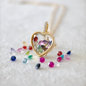 Può includere: Una collana con pendente a forma di cuore dorato, con un centro trasparente pieno di pietre preziose colorate. Il pendente è impreziosito da piccole pietre trasparenti ed è appeso a una delicata catena d'oro. Varie pietre preziose sciolte rosse, blu, verdi e rosa sono sparse.