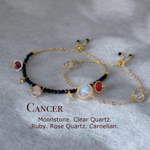 May include: Two gold bracelets with black beads and gemstone charms. The charms include a moonstone, clear quartz, ruby, rose quartz, and carnelian. The bracelets are adjustable with a sliding knot. The text "CANCER" is written below the bracelets, along with the names of the gemstones.