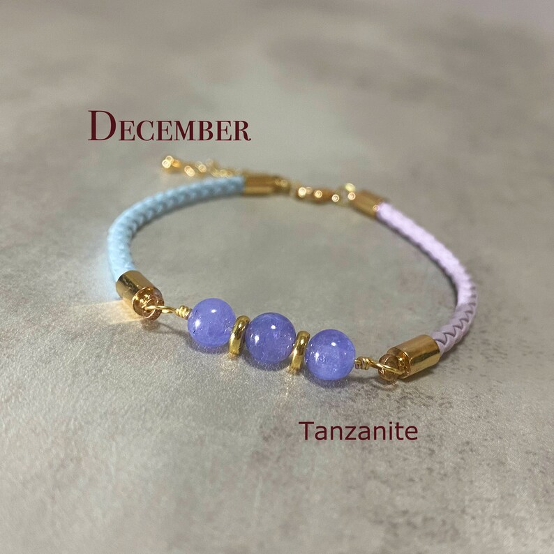 May include: A light blue and lavender braided cord bracelet with three blue Tanzanite beads and gold accents. The bracelet has the word "DECEMBER" written above it and the word "Tanzanite" written below it.