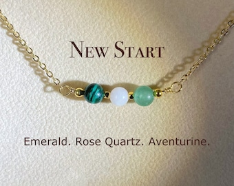 May include: A gold chain necklace with four beads: green malachite, white rose quartz, green aventurine, and a gold bead. The necklace has the text "NEW START" above the beads and the text "Emerald. Rose Quartz. Aventurine." below the beads.