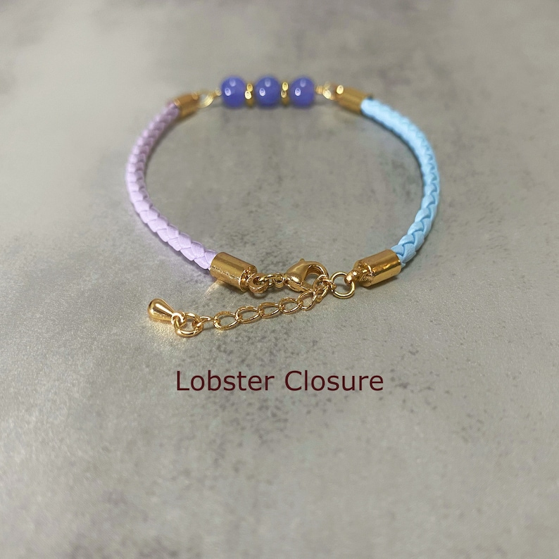 May include: A light blue and lavender braided leather bracelet with three blue gemstone beads and a gold lobster clasp closure.
