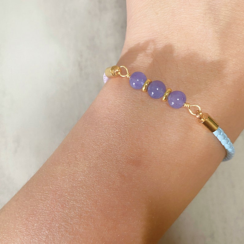 May include: A light blue and purple braided leather bracelet with three purple gemstone beads and gold accents.