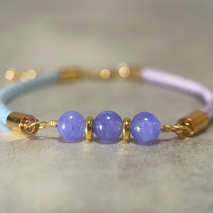 May include: A light blue and pink braided bracelet with three purple gemstone beads and gold accents.