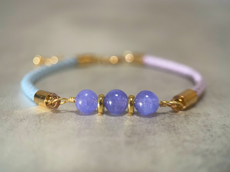 May include: A light blue and pink braided bracelet with three purple gemstone beads and gold accents.