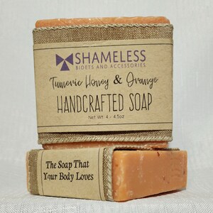May include: Two bars of orange-colored handmade soap with a brown paper label that reads "SHAMELESS BIDETS AND ACCESSORIES Turmeric Honey & Orange HANDCRAFTED SOAP Net Wt. 4 - 4.5oz" and a second label that reads "The Soap That Your Body Loves".