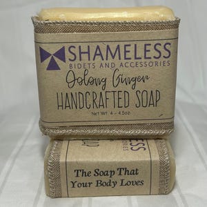 May include: Two bars of handcrafted soap. The top bar is wrapped in a brown paper label with the text "SHAMELESS BIDETS AND ACCESSORIES Oolong Ginger HANDCRAFTED SOAP Net Wt. 4 - 4.5oz." The bottom bar reads "The Soap That Your Body Loves."