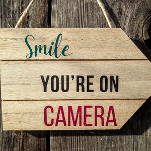 May include: Wooden arrow-shaped sign with the words "Smile YOU'RE ON CAMERA" in teal, black, and red. The sign is made of light-colored wood and hangs from a natural twine string. The background is a weathered wooden wall.
