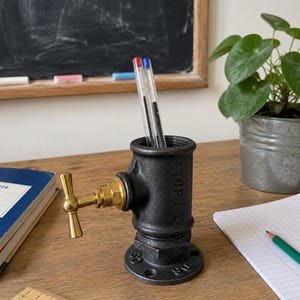 Peut inclure: Un porte-crayon en raccord de tuyau noir avec une vanne à poignée en T couleur laiton. La base cylindrique présente une bride circulaire avec des trous de boulons. Deux stylos sont insérés dans l'ouverture supérieure. Une règle en bois est visible.