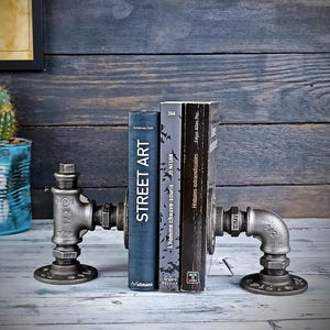 Può includere: Reggilibri in stile industriale realizzati con tubi metallici, che sostengono due libri. Uno è intitolato "STREET ART", l'altro ha testo in francese. I reggilibri hanno una finitura metallica scura e sono su uno sfondo di legno rustico.