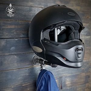Może przedstawiać: Czarny kask motocyklowy marki Bell zamontowany na metalowym wsporniku rurowym, z niebieską kurtką wiszącą poniżej. Wspornik jest przymocowany do ściany z ciemnymi panelami drewnianymi. Obraz zawiera logo w lewym g&oacute;rnym rogu.