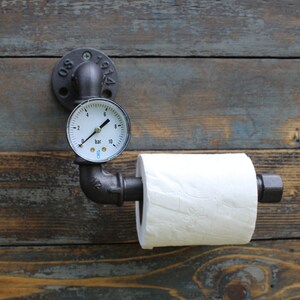 May include: Industrial-style toilet paper holder made from black metal pipes and fittings. Features a pressure gauge and a roll of white toilet paper. Mounted on a weathered wooden wall. The gauge reads in bar units.