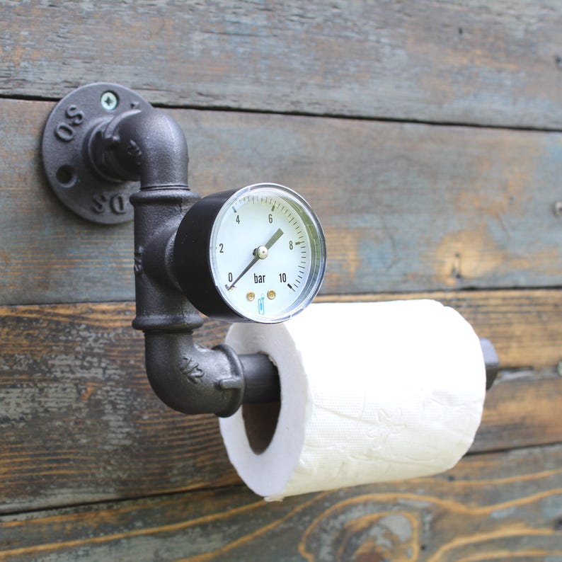 May include: Industrial-style black metal toilet paper holder mounted on a wooden wall. The holder incorporates a pressure gauge and pipe fittings, with a roll of white toilet paper in place. The gauge reads in bar.