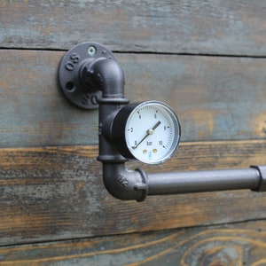 May include: Industrial-style toilet paper holder made from black metal pipes. Features a pressure gauge with a white face and black markings. Mounted on a weathered wood background.