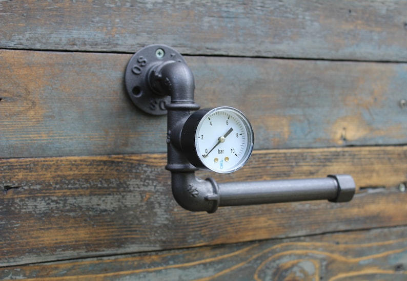 May include: Industrial-style toilet paper holder made from black metal pipes. Features a pressure gauge with a white face and black markings. Mounted on a weathered wood background.