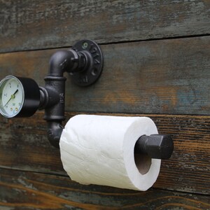 May include: A black industrial-style toilet paper holder mounted on a weathered wooden wall. The holder features a pressure gauge and a roll of white toilet paper. The design incorporates black pipes and fittings, creating a unique, rustic aesthetic.