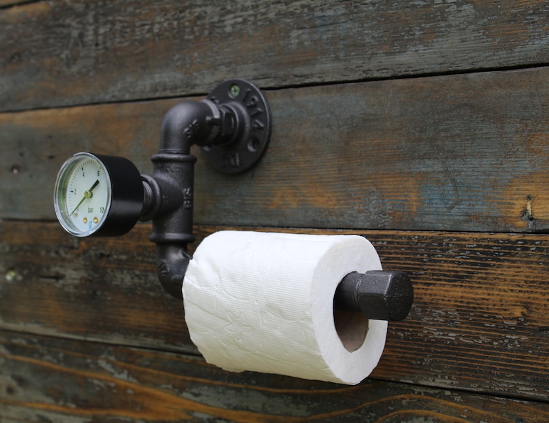 May include: A black industrial-style toilet paper holder mounted on a weathered wooden wall. The holder features a pressure gauge and a roll of white toilet paper. The design incorporates black pipes and fittings, creating a unique, rustic aesthetic.