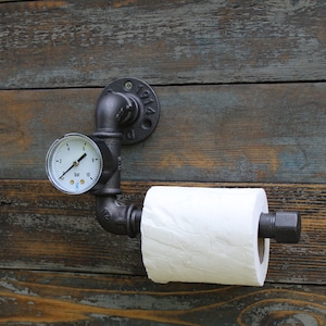 May include: Industrial-style toilet paper holder made from black metal pipes, mounted on a rustic wooden wall. Features a pressure gauge and a roll of white toilet paper. The metal fixture has the year "1914" stamped on it.