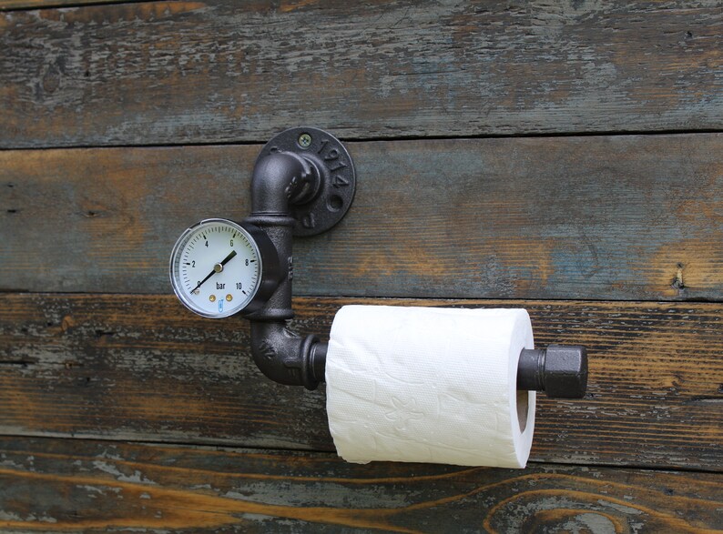 May include: Industrial-style toilet paper holder made from black metal pipes, mounted on a rustic wooden wall. Features a pressure gauge and a roll of white toilet paper. The metal fixture has the year "1914" stamped on it.