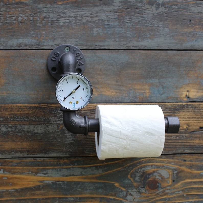 May include: Industrial-style toilet paper holder made from black metal pipes and fittings. Features a pressure gauge and a roll of white toilet paper. Mounted on a weathered wooden wall. The gauge reads in bar units.