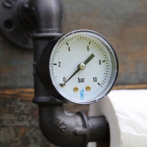 May include: Industrial-style toilet paper holder featuring a pressure gauge with a white face and black markings, reading in bar units. The gauge is mounted on black metal pipes, with a roll of white toilet paper visible.
