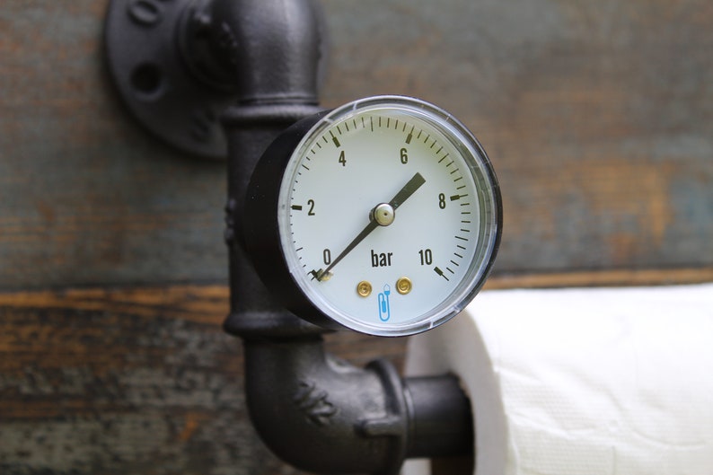 May include: Industrial-style toilet paper holder featuring a pressure gauge with a white face and black markings, reading in bar units. The gauge is mounted on black metal pipes, with a roll of white toilet paper visible.
