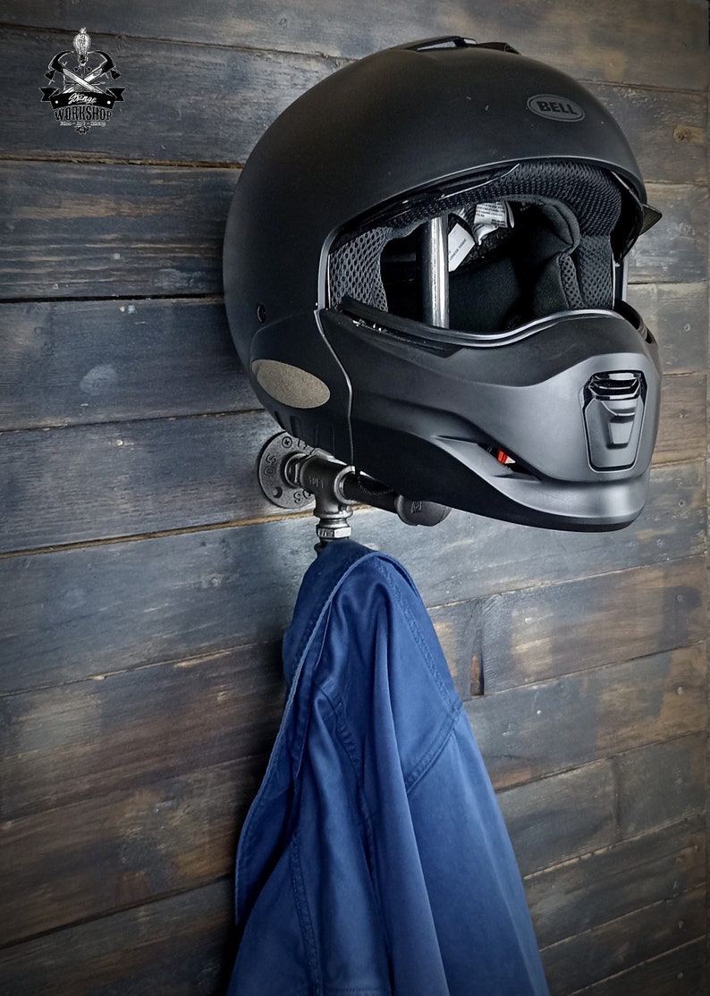 Może przedstawiać: Czarny kask motocyklowy marki Bell zamontowany na metalowym wsporniku rurowym, z niebieską kurtką wiszącą poniżej. Wspornik jest przymocowany do ściany z ciemnymi panelami drewnianymi. Obraz zawiera logo w lewym g&oacute;rnym rogu.