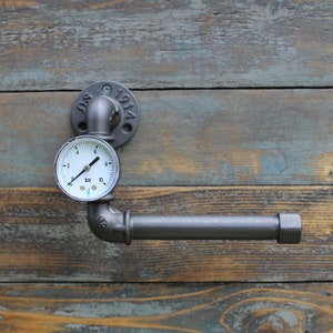 May include: Industrial-style toilet paper holder made from dark gray metal pipes. A pressure gauge is mounted above the horizontal bar. The holder is attached to a wooden surface. The flange is stamped with "SO 1914".