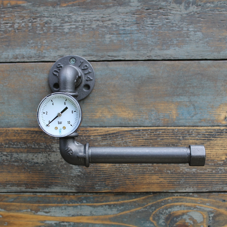 May include: Industrial-style toilet paper holder made from dark gray metal pipes. A pressure gauge is mounted above the horizontal bar. The holder is attached to a wooden surface. The flange is stamped with "SO 1914".
