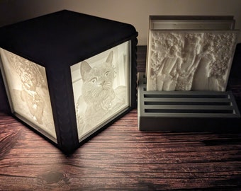 May include: A black, cube-shaped lamp with illuminated panels displaying a cat portrait and other images. Beside it, a gray holder contains several white relief tiles depicting a scene with figures and trees. The items are on a wooden surface.