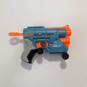 Puede incluir: Un blaster de juguete Nerf Elite 2.0 Volt azul y naranja con una empuñadura negra y una manija de cebado naranja. El blaster tiene un cañón negro con un diseño de malla y el texto "VOLT" impreso en el lateral.