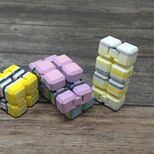 May include: Three colourful fidget toys made of soft, textured plastic. The toys are shaped like cubes and have different patterns and colours, including yellow, pink, and black.