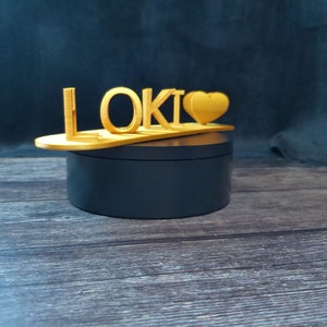 May include: A wooden sign with the word "LOKI" and a heart shape, sitting on a black round base.