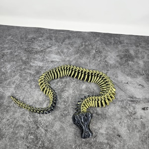 May include: A segmented, articulated snake toy in black and gold. The snake's body is made of connected segments, with a detailed, textured appearance. The head is a darker, solid black, and the body has a metallic gold finish.