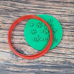 Könnte beinhalten: Ein grüner Plastik-Ausstecher mit den Worten "Merry & Bright" und Schneeflocken. Der Ausstecher liegt auf einer Holzoberfläche.