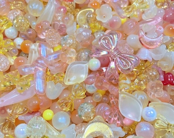 May include: A close-up shot of a mix of colorful beads in various shapes and sizes. The beads are in shades of pink, yellow, and clear, with some iridescent and pearlized finishes. The assortment includes bows, flowers, and other decorative elements.