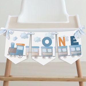 Könnte beinhalten: Ein Banner mit einem Zugdesign und dem Wort "ONE" in Blau und Orange. Das Banner ist weiß mit einem blauen Zug- und Wolkenmuster. Es ist mit blauen Schleifen an einem Hochstuhl befestigt.