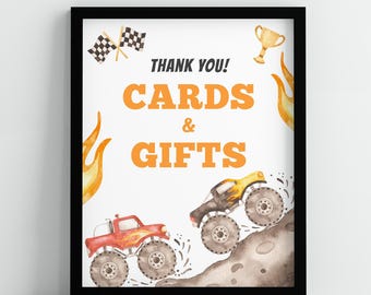 May include: A white sign with black text that says "Thank You! Cards & Gifts". The sign is decorated with two monster trucks, one red and one yellow, driving on a dirt road. There are flames on either side of the sign and a checkered flag and a gold trophy at the top.