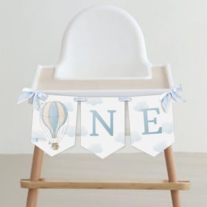 May include: A blue and white hot air balloon high chair banner with the word "ONE" in blue letters. The banner is decorated with white clouds and blue ribbons.