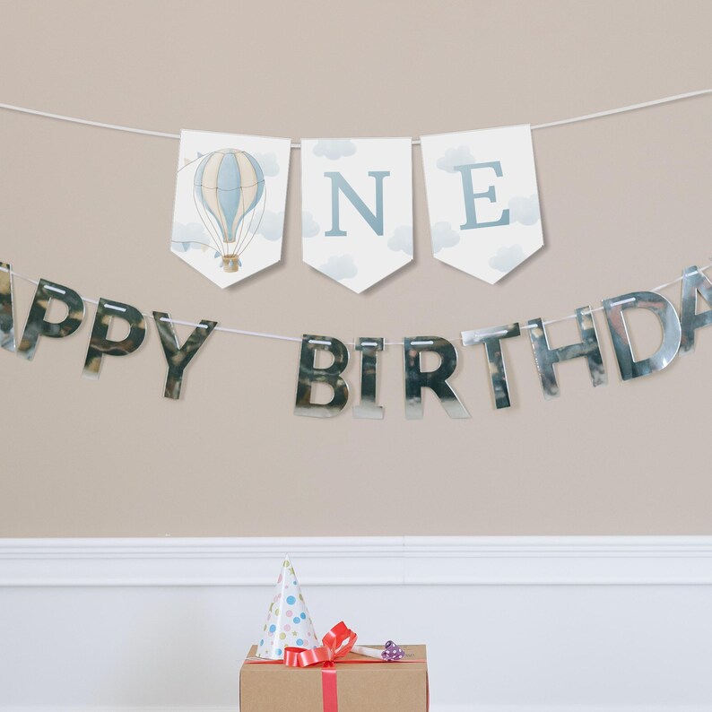 May include: A silver Happy Birthday banner with a blue and white banner that says "ONE" with a hot air balloon graphic.